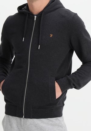 Hombre con sudadera negra con cremallera y manos en los bolsillos, pequeña "F" naranja bordada en el pecho y pantalones deportivos grises.