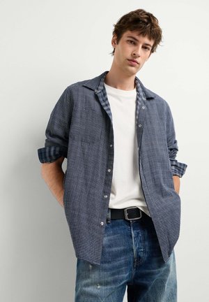 Junger Mann mit den Händen in den Taschen, trägt ein weißes T-Shirt, ein blau kariertes Hemd und blaue Jeans vor einem einfarbigen hellen Hintergrund.
