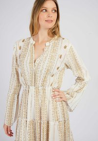 Langärmliges Kleid aus leichtem Stoff, mit einem cremefarbenen Hintergrund und goldenen Blumen- und geometrischen Mustern. Knopf-Verschluss am Ausschnitt, gestuftes Design.