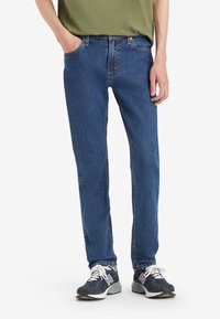 Personne portant un jean denim bleu, des baskets noires et blanches, et une chemise vert olive avec une main dans la poche sur un fond uni.