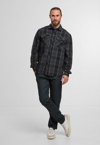 Schwarz-graues kariertes Button-Up-Hemd mit zwei Brusttaschen, kombiniert mit dunklen Hosen und hellen Sneakers vor einem neutralen Hintergrund.