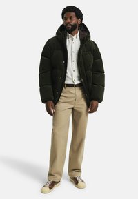 Doudoune vert sombre avec capuche, présentant un design matelassé. Portée par-dessus une chemise claire, assortie à un pantalon beige et des chaussures marron.