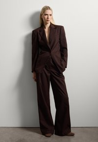 Bruin op maat gemaakt blazer met brede schouders, gecombineerd met hoogstaande, wijde broekspijpen. Glad stof met minimale details.