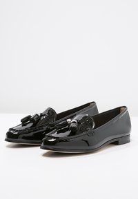 Mocassins en cuir verni noir avec des franges, à talons bas et bouts arrondis, placés côte à côte sur une surface blanche.