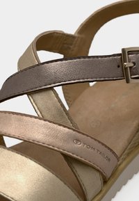 Κοντινό πλάνο μεταλλικού χρυσού και μπρούτζινου σανδαλιού με λουράκια, αγκράφα και το λογότυπο "Tom Tailor" σε ένα από τα λουράκια.