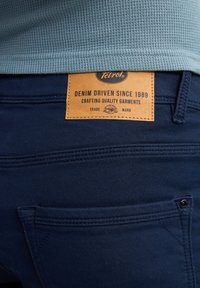 Jeans de mezclilla azul marino con una etiqueta de cuero que muestra el logo "Tetrol". Las costuras visibles y los detalles de los bolsillos realzan el diseño.