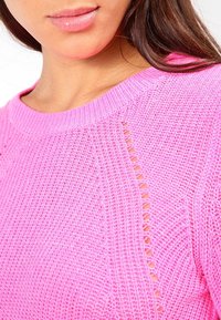 Rosa Strickpullover mit strukturiertem Muster, das horizontale und vertikale Streifen sowie kleine Lochdetails an den Seiten aufweist.