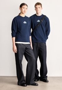 Tengerészkék pulóverek logóval, sötét farmer széles szárú nadrággal párosítva. A férfi modell oldaltáskával viseli. Mindenütt sima textúrák.