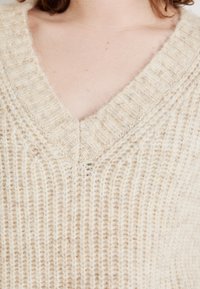 Beige kötött pulóver V nyakkal, bordás textúrával és puha szálakkal, hangsúlyozva a melegséget és kényelmet az anyagban.