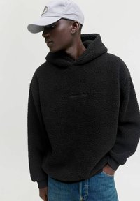 Schwarze Fleece-Hoodie mit einer strukturierten, flauschigen Oberfläche und einer Kängurutasche. Kombiniert mit blauen Jeans und einer hellgrauen Mütze.