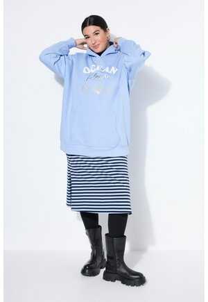 Femme portant un sweat à capuche oversize bleu clair avec le texte "OCEAN please" et des motifs de coquillages, jupe rayée, leggings noirs et bottes noires.