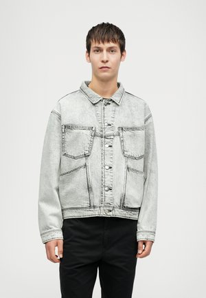 Ung mand iført en lys grå vaskede denimjakke med store frontlommer og sorte bukser, stående mod en simpel hvid baggrund.