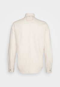 Camicia a maniche lunghe color crema vista dal retro con colletto alto, cucitura centrale e polsini abbottonati.
