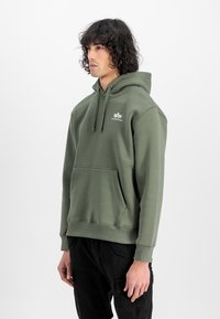 Olivgrüner Hoodie aus weichem Stoff, mit einer Kängurutasche, Zugbandkapuze und kleinem weißen Logo auf der Brust. Klassische Passform mit gerippten Bündchen.