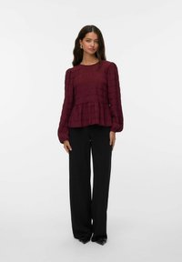 Blouse bordeaux texturée avec de longues manches bouffantes et un ourlet à volants, associée à un pantalon noir à jambes larges et des escarpins noirs pointus.
