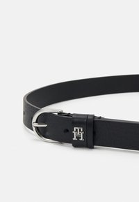 Curea din piele neagră cu o cataramă din metal argintiu. Are o textură netedă și o buclă din piele cu marcă. Design simplu și elegant, cu detalii minimale.