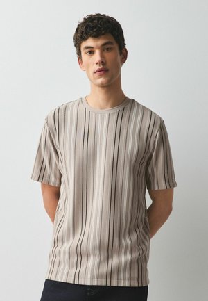 Ung mand med kort krøllet hår iført en beige kortærmet skjorte med lodrette sorte og hvide striber, stående mod en ensartet lys baggrund.