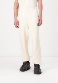 Cremefarvede sweatpants med en afslappet pasform og et broderet grønt logo på den ene side. Sammen med mørke mønstrede sneakers.