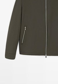 Olivengrøn jakkeærme og nederste del af torsosektionen med sølvfarvede lynlåse foran og på siden, vist på hvid baggrund.