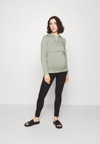 Sweatshirt de maternité vert avec le texte "MAMA", manches longues et une couche avant. Associé à desleggings noirs et des sandales à enfiler noires.