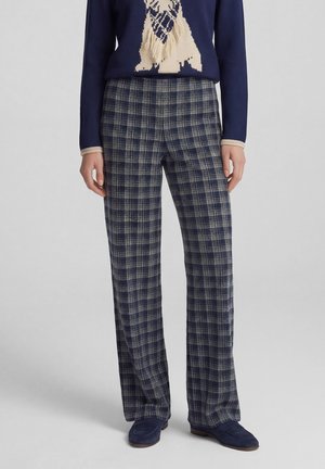 Navy plaid straight-leg trousers worn with navy loafers and a navy sweater with beige fringe detail at front.