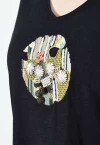 Zwarte v-hals t-shirt met intricate, multifunctionele borduursels van schelpen, bloemenpatronen en diverse texturen op de voorkant.