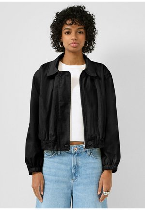 Kobieta o kręconych włosach ubrana w czarną kurtkę na zamek, białą koszulę, jasnoniebieskie dżinsy, srebrne kolczyki koła oraz wiele srebrnych pierścionków, stoi na tle białej ściany.