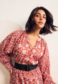 Czerwona sukienka w kwiaty z białymi wzorami, z dekoltem w serek, długimi rękawami i czarnym skórzanym paskiem. Materiał o strukturze z detalicznym przeszyciem.