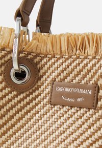 Torebka z plecionego słomy w kolorze beżowym z postrzępioną górą, brązowymi skórzanymi akcentami, metalowym pierścieniem oraz markową skórzaną metką z napisem "Emporio Armani Milano 1981."
