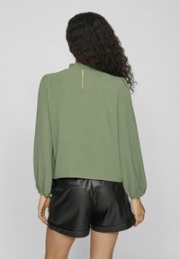 Groene, gestructureerde blouse met lange ballonmouwen, voorzien van een opening met sleutelhoed aan de achterkant, gecombineerd met zwarte leren shorts.