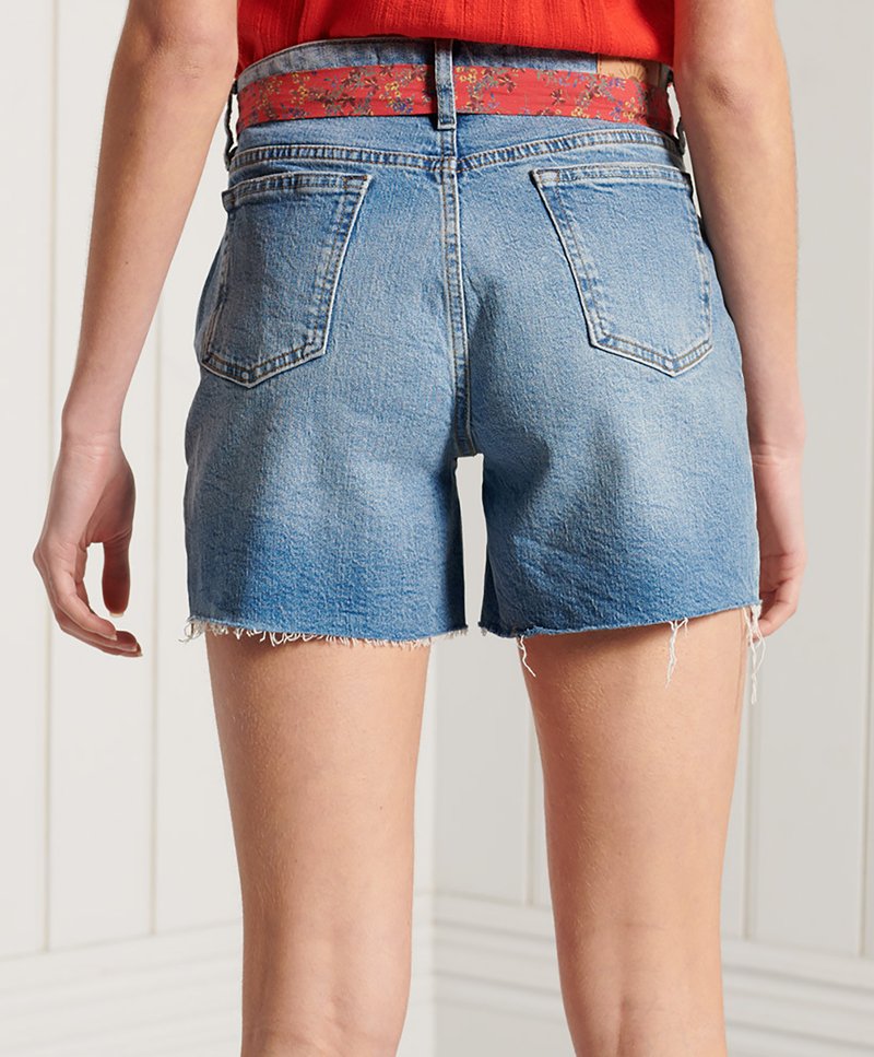 Rückansicht einer Person in hellblauen Jeansshorts mit ausgefransten Säumen und einem roten Blumen-Gürtel, die vor einer weißen Holzpaneelwand steht.