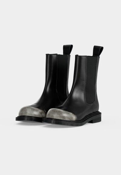 Paire de bottes en cuir noires montant mi-mollet avec embouts renforcés argentés métalliques et languettes de tirage sur un fond clair uni.