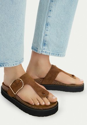 Füße in hellblauen Jeans, die braune Wildledersandalen mit breitem Riemen, goldener Schnalle und dicker schwarzer Sohle tragen.