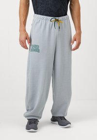 Světlé šedé tepláky s elastickým pasem, zelenožlutou šňůrkou na stažení a grafikou "Under Armour" v zelené barvě na levé nohavici.