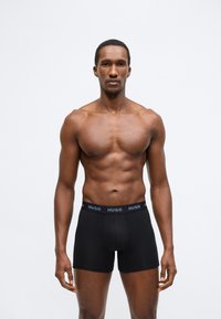 Melni bokseršorti ar zilu logotipa jostu, izgatavoti no elastīga auduma. Modelis stāv taisni, demonstrējot pieguļošu dizainu.