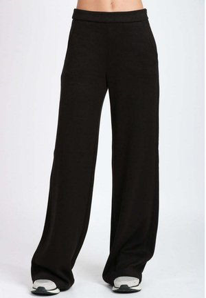 Persona che indossa pantaloni neri larghi e scarpe da ginnastica bianche, in piedi su uno sfondo chiaro e semplice, visibile dalla vita ai piedi.