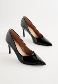 Zwarte lakleren pumps met een puntige neus, een slanke hak en een glad oppervlak. De binnenkant is voorzien van een beige voering.