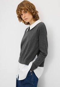 Grauer Pullover mit weißem Kragen, über einem weißen Hemd getragen. Mit langen Ärmeln und glatter Textur. Kombiniert mit blauen Jeans.