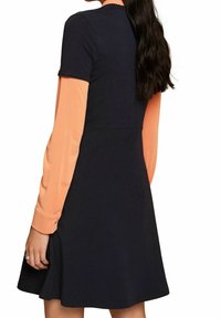 Granatowa sukienka z krótkimi rękawami i rozkloszowanym dołem, nałożona na jasnopomarańczową bluzkę z długim rękawem. Miękka tkanina, prosty krój, bez wzorów.