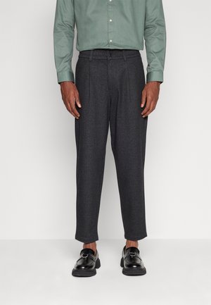 Pantaloni grigi texture con vestibilità rilassata, caratterizzati da pieghe e una vita elastica, abbinati a mocassini neri e una camicia verde con bottoni.