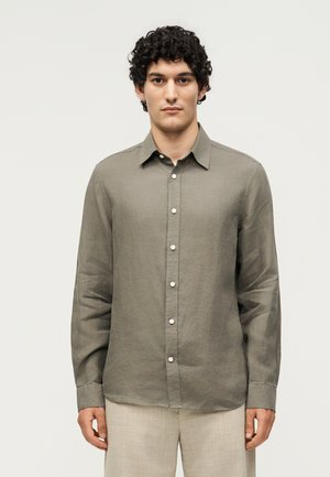 Młody mężczyzna z kręconymi ciemnymi włosami, ubrany w zapinaną na guziki oliwkowo-zieloną koszulę z długim rękawem i jasnobeżowe spodnie, stojący na jednolitym tle.