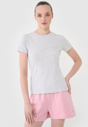 Młoda kobieta z włosami związanymi do tyłu, ubrana w jasnoszary t-shirt z krótkim rękawem i różowe krótkie spodenki, stoi z jedną ręką na biodrze, a drugą luźno.