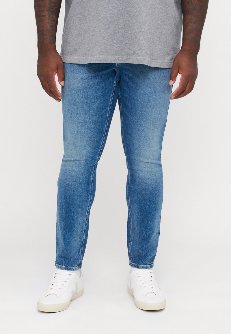 Blugi din denim bleu deschis, cu croială slim, prezentând o uzură subtilă și cusături în contrast. Asortați cu adidași albi.