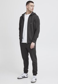 Grijze zip-up hoodie met een kangoeroezak, gecombineerd met bijpassende joggingbroek en een wit T-shirt. Sneakers met rubberen zolen maken de look compleet.