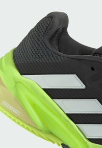 Zapatilla deportiva negra con parte superior texturizada, acentos de suela en verde neón y tres franjas plateadas a lo largo del costado. Incluye la etiqueta "REPETITOR".
