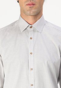 Light gray button-up shirt with a textured pattern, featuring a classic collar and brown buttons. Neutral color suitable for various occasions.
