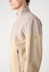 Casaco bege claro com uma secção superior bege estampada, gola alta e fecho lateral. Apresenta um logotipo na manga e uma textura suave.