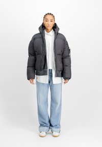 Vrouw met gevlochten haar in een zwart pufferjack, witte overhemd, lichtblauwe wijde spijkerbroek en witte sneakers staat tegen een witte achtergrond.