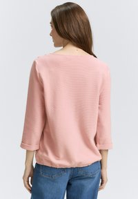 Helles rosa Bluse mit Dreiviertelärmeln, rundem Ausschnitt, strukturiertem Stoff und umgeschlagenen Manschetten. Der Rücken ist mit dezenten Knopfdetails versehen.
