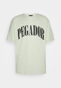 Vaaleanvihreä puuvillainen T-paita, jossa on lyhyet hihat ja pyöreä pääntie. Paidassa on suuret mustat kirjaimet, jotka muodostavat kaarevan "PEGADOR"-tekstin.